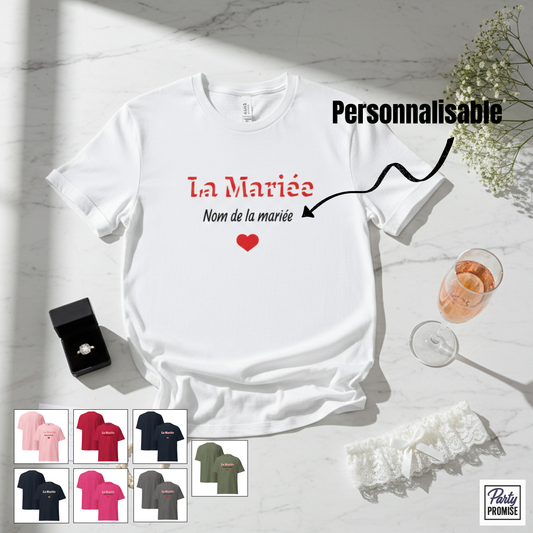 T-shirt blanc personnalisable “La Mariée” avec nom de la mariée et petit cœur rouge, présenté en mise en scène EVJF avec bague, coupe de champagne et jarretière, disponible en plusieurs couleurs pour enterrement de vie de jeune fille.