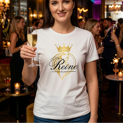 Femme souriante lors d'une soirée festive, portant le t-shirt "Reine du jour" et tenant une coupe de champagne dans un salon chic.